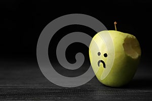 Bitten apple with drawing of unhappy face on table against dark background, space for text.