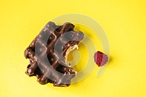 Bitten off chocolate waffle and raspberry, yellow background, top view