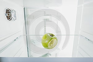 Bitten off green apple on shelf of refrigerator with open door isolated on white