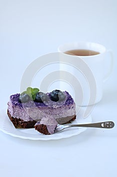 Bitten off purple blueberry mousse cake with blueberry on white plate with teaspoon and cup of tea on white background