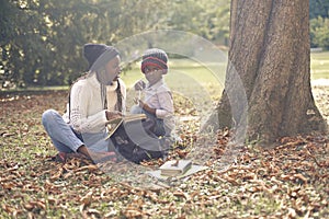 Black adult woman with long braids reading a book with a young child in a park