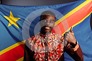 A black African man smiling, posing, showing thumb UP in front of the flag of the Democratic Republic of the Congo.