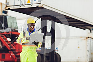 Black African worker working in logistic shipping using laptop to control loading containers at port cargo for import export goods