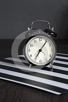 Black alarm clock on a black and white striped napkin showing 7 o`clock on a bedside table