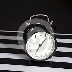 Black alarm clock on a black and white striped napkin showing 7 o`clock on a bedside table
