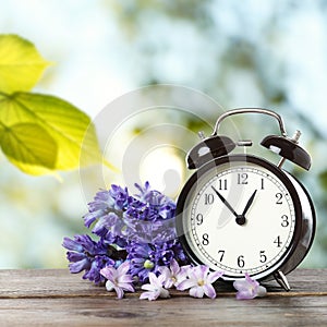 Black alarm clock and flowers on wooden table against blurred background. Spring time