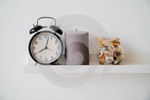 Black alarm clock, jar with dried flowers and perfume and gray candle on shelf.