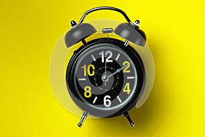 A black alarm clock with large numbers lies on a yellow background. The concept of time or deadline.