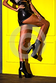 Black ankle boots on a high platform and high heels leather on a model that stands in a black jacket and black shorts on a yellow
