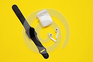 Black AppleWatch and white AirPods with box on bright yellow background. Isolated, copy space for text or advertisement.