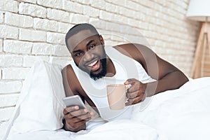 Black, awakened man drinks coffee in bed browsing