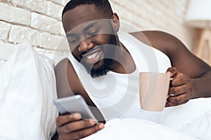 Black, awakened man drinks coffee in bed browsing