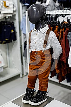A black baby mannequin in a shirt, suspender pants, and black shoes in a shopping center. Children`s casual clothing store. Moder