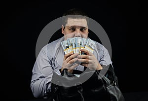 On a black background, a man in a business shirt holds a fan of stacks of hundred dollar bills in front of his face
