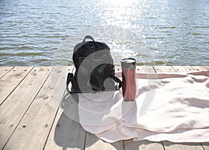 Black backpack and red thermos with hot tea on plaid on a wooden pier against background of the Japanese sea and sun rays. travel