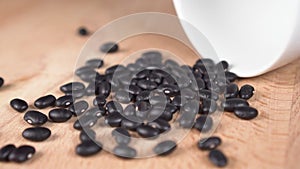 Black beans spilling from a white ceramic bowl in slow motion onto a wooden surface.