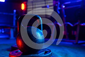 Black big kettlebell in the gym with red and blue light. An old round kettlebell with a handle and a barbell rack in the