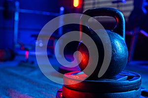 Black big kettlebell in the gym with red and blue light. An old round kettlebell with a handle and a barbell rack in the