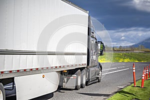 Black big rig bonnet semi truck transporting cargo in dry van semi trailer with aerodynamic skirt running on the local road with