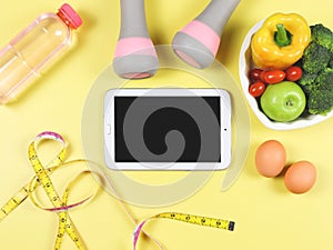 Black blank screen computer tablet , vegetables and fruit in heart shape plate, bottle of water, measuring tape,  pink dumbbells