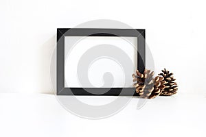 Black blank wooden frame mockup with pine cones lying on the white table. Poster product design. Styled stock feminine