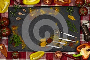 Black board with scattered spices, iron spoons, fresh vegetables on a red and white cloth background