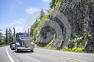 Black bonnet classic big rig semi truck tractor transporting cargo on flat bed semi trailer driving on the winding mountain road