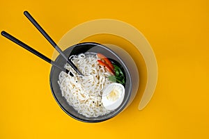 Black bowl of chineese fast cooked noodle with egg and vegetables. Black chopsticks near it at yellow background. photo