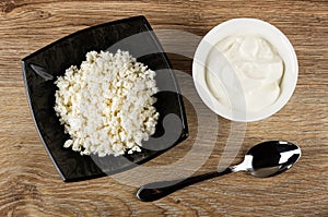 Black bowl with defatted cottage cheese, bowl with sour cream, spoon on wooden table. Top view