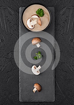 Black bowl plate of creamy chestnut champignon mushroom soup on black table background with black stone board and fresh mushrooms