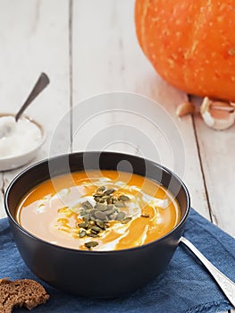 Black bowl with pumpkin soup, sauce and pumpkin seeds on a blue linen napkin. Sauce, pumpkin and garlic in the background. Close-