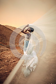 Black bride stands and holds waving bridal veil in her hands on background of beautiful landscape.