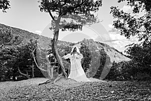 A black bride in a white dress stands near an old tree against the backdrop of mountains, black and white version