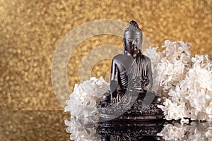 Black Buddha framed by white lilac flowers on a golden bokeh background with reflection