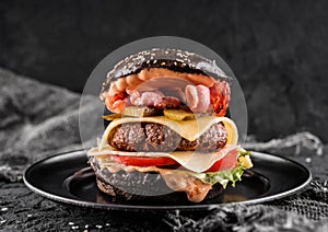 Black burger with beaf steak, lettuce, tomatoes, slice of cheese, ham, pastrami and sauce on slate black background, close up