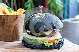 Black burger with meat patty, cheese, tomatoes, mayonnaise, french fries in a wooden plate. Modern fast food lunch.