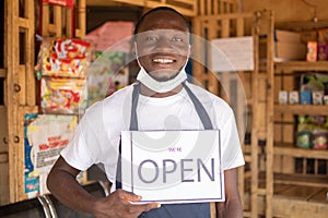 Black business owner wearing a face mask and holding an open sign in front of his shop and smiling