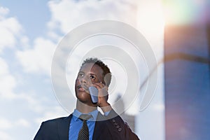 black businessman with blue suit and tie using mobile, calling, working outside building in city with sunrise or sunset light.