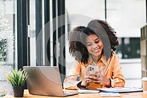 Black businesswoman using smartphone while working on a laptop computer. Smiling African American woman using mobile