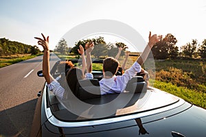 Black cabriolet is on the country road. Company of young girls and guys are sitting in the car hold their hands up on a