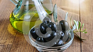 Black canned olives in glass transparent bowl, olive oil bottle in sunlight on the wooden table. Healthy mediterranean food