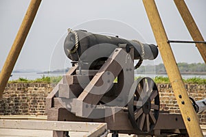 A black cannon with a brown wooden base in front of a red brick wall with lush green plants along the Savannah River