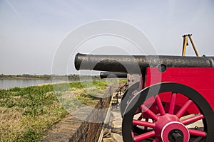 black cannons with a brown and red wooden base in front of a red brick wall with lush green plants and trees along the Savannah