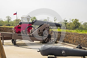 black cannons with a brown wooden base in front of a red brick wall with lush green plants and trees along the Savannah River