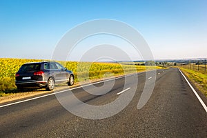 Black car parked on road side with field of golden sunflower background .