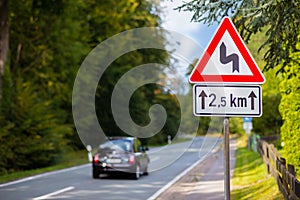 A black car is racing. A warning sign in front of a very winding country road.