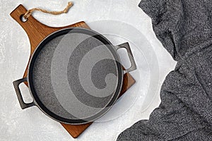 Black cast iron empty frying pan on a wooden cutting board and a gray kitchen towel on a gray concrete background