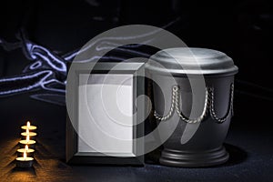 Black cemetery urn with black blank mourning frame and burning candles, on deep blue background