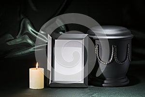 Black cemetery urn with black mourning frame, burning candle, on deep green background