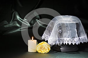 Black cemetery urn with burning candle, white lace shroud, yellow rose on deep green background